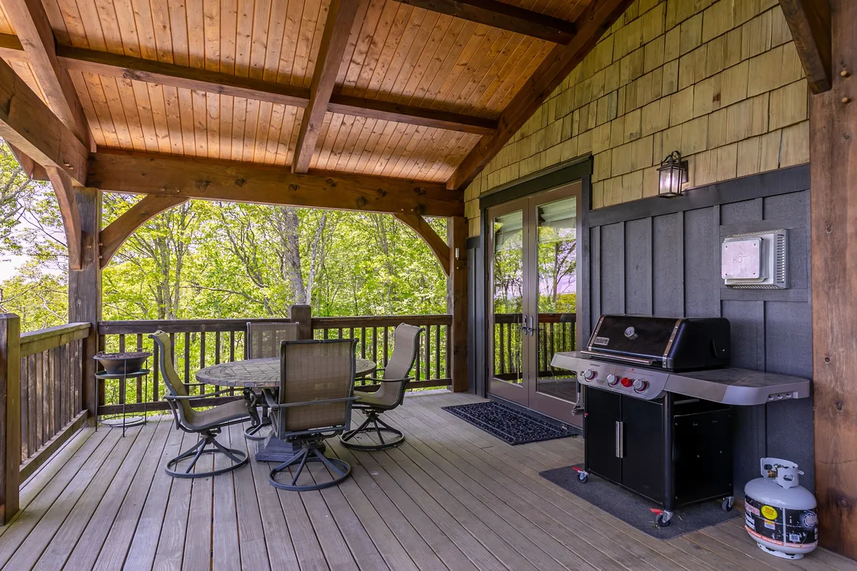 A cozy exterior dining area with a grill, a wooden dining table, and surrounding forest greenery. The atmosphere is inviting, with hints of sunset or early evening, perfect for barbecues, group gatherings, and enjoying nature just steps outside the home.