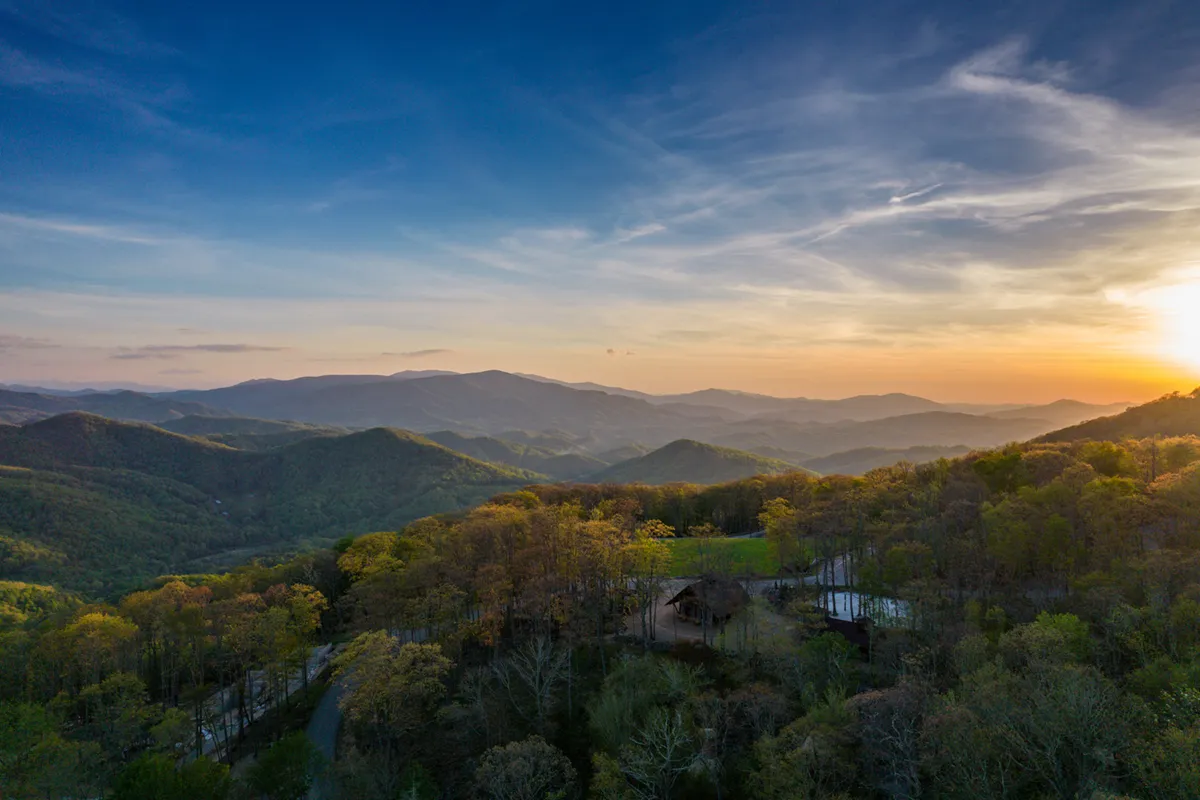 The scene highlights a panoramic mountain range at sunset, with a forested landscape and a glimpse of the property structure in the foreground. The colors of the sky and the sweeping view emphasize the majesty of the Blue Ridge setting.
