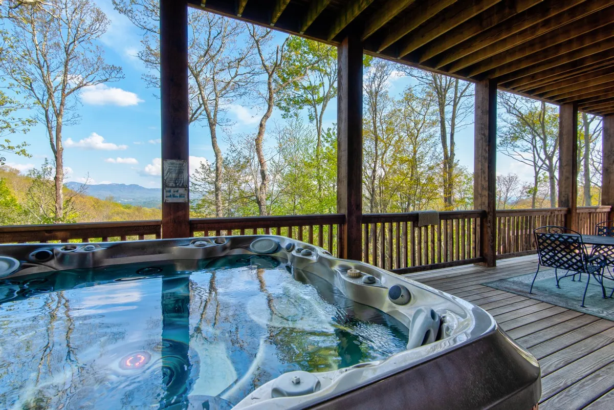 A wooden deck features a hot tub overlooking dramatic mountain vistas, surrounded by trees. The setting is serene, ideal for family relaxation after a day of adventure, with the spacious deck suggesting group gatherings and relaxing moments together.