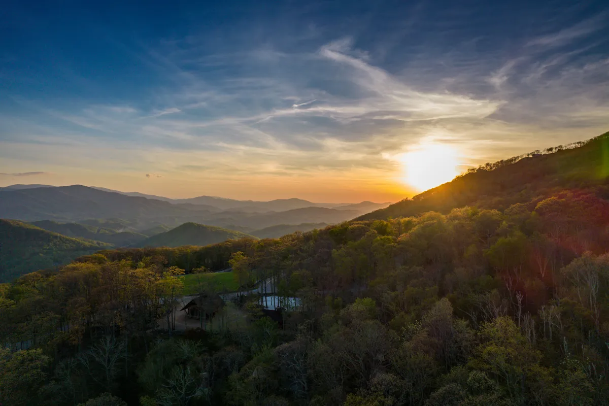 A sweeping vista captures a mountain range at sunset, dense woods below, and a cozy cabin nestled in the scene. The landscape suggests limitless opportunities for hiking, exploring trails, and enjoying the serenity of nature.