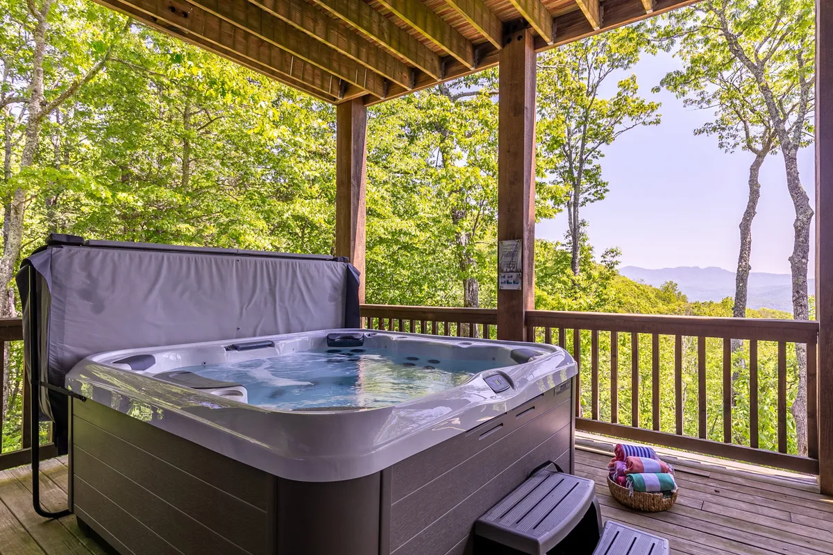 A tranquil outdoor hot tub sits on a wooden deck surrounded by tall trees, with an expansive view of the Blue Ridge Mountains in the background. The setting exudes peace and relaxation after a day of outdoor adventure, perfectly blending natural beauty with comfort.