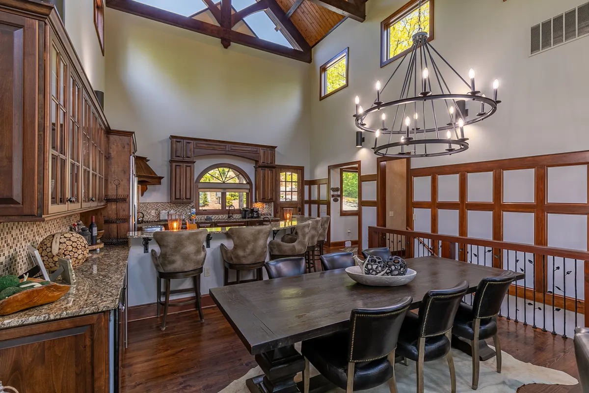 A spacious, welcoming dining area with a large dining table set under a chandelier, adjacent to a kitchen bar. Exposed wooden beams and large windows create a cozy yet elegant atmosphere, inviting gatherings, celebrations, and shared meals.