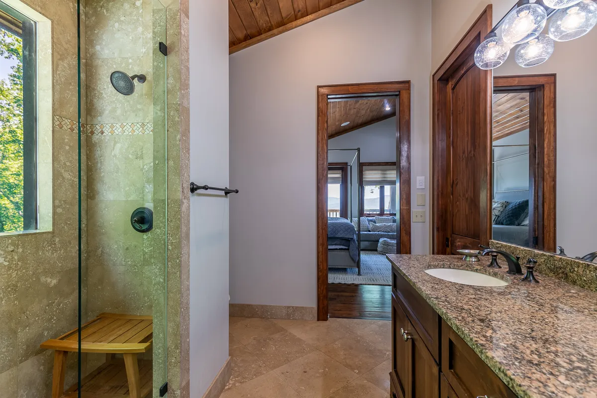 A spa-inspired bathroom featuring a walk-in glass shower with a rainfall showerhead, granite countertop vanity, a wooden bench, and a framed mirror. The ambiance is both tranquil and luxurious, embodying self-care and relaxation.