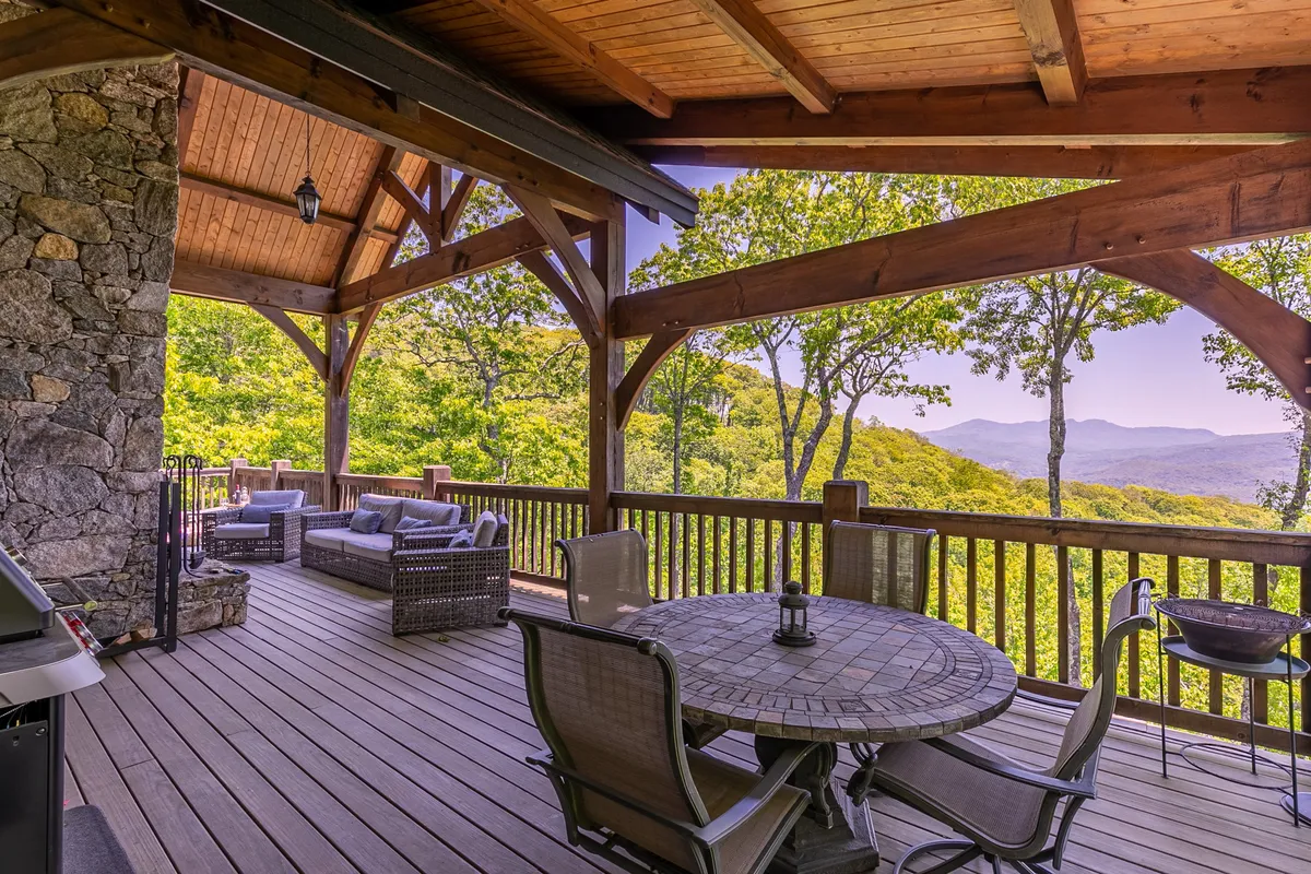 A spacious exterior patio featuring a wooden dining table set beneath timber beams, with outdoor seating and a breathtaking view of the mountains beyond. The stone wall and open layout create a perfect spot for group dining or social gatherings against a scenic natural backdrop.
