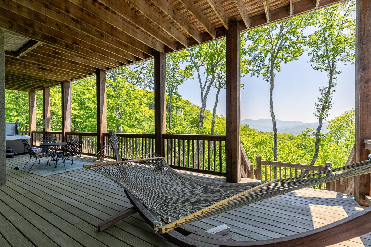 An inviting deck with a hammock, outdoor dining table, and chairs surrounded by trees and views of distant mountains. The setting suggests relaxation, group meals, and enjoying nature together.
