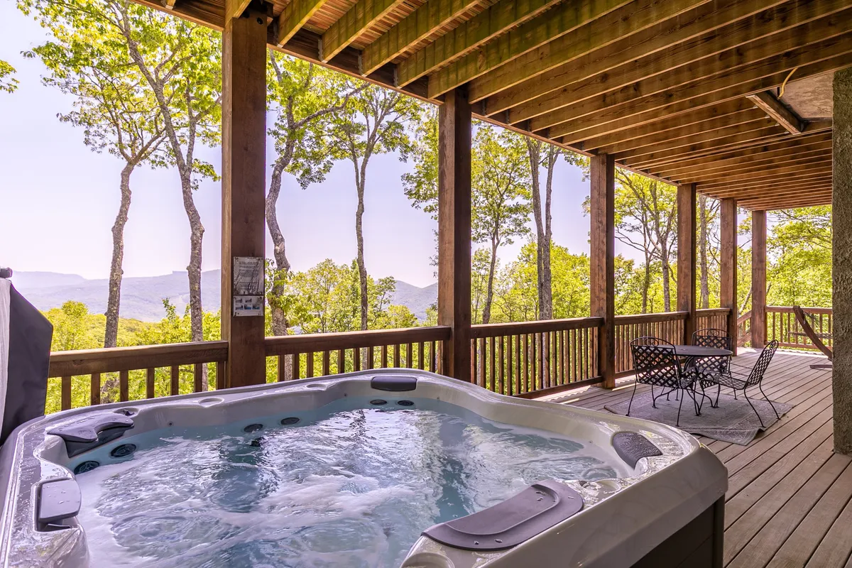 A spacious wooden deck features a bubbling hot tub, comfortable outdoor seating, and a panoramic view of the mountains. The area invites guests to relax, socialize, and enjoy the outdoors, day or night.