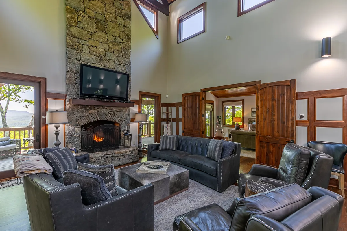 A cozy great room featuring a prominent stone fireplace, comfortable seating, and floor-to-ceiling windows that frame expansive mountain views. The natural wood finishes and ample light exude warmth and relaxation.
