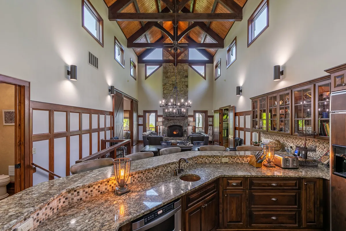 A breathtaking great room anchored by a massive stone fireplace, soaring vaulted timber ceilings, a grand chandelier, and an open-plan layout that flows seamlessly into a modern kitchen. Sunlight streams through expansive windows, illuminating the high-end finishes and inviting gathering spaces.